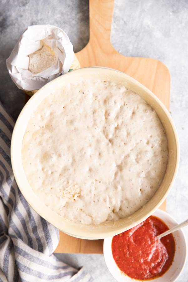 Schüssel mit selbstgerechtem Pizzateig, daneben eine Schale mit Tomatensauce und eine Packung Mehl