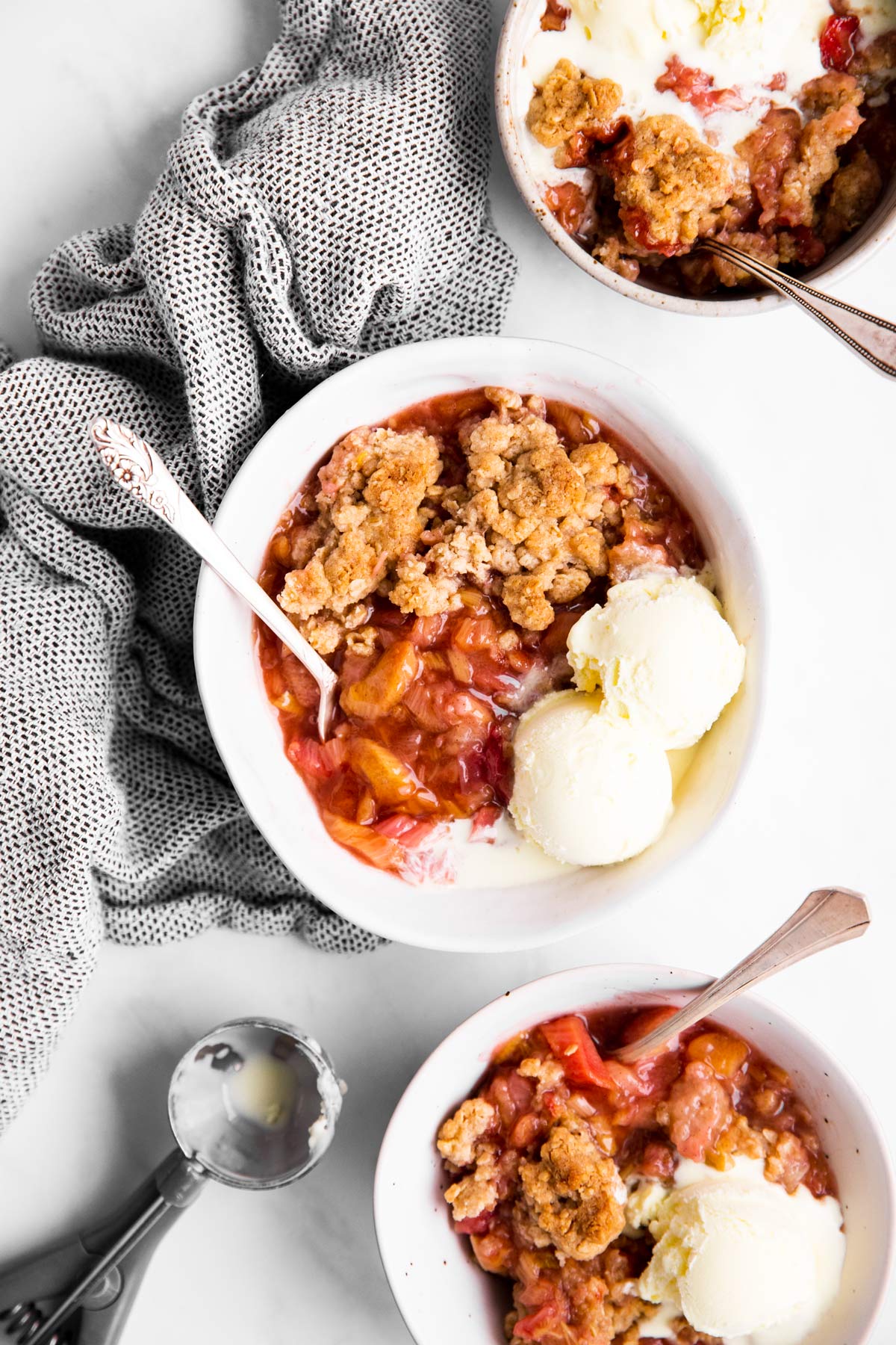 drei weisse Schalen mit Rhabarbercrumble und Vanille-Eis auf einer hellen Oberfläche